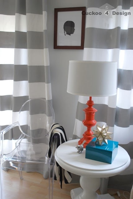 orange painted brass lamp on white side table with horizontal striped gray and white curtains