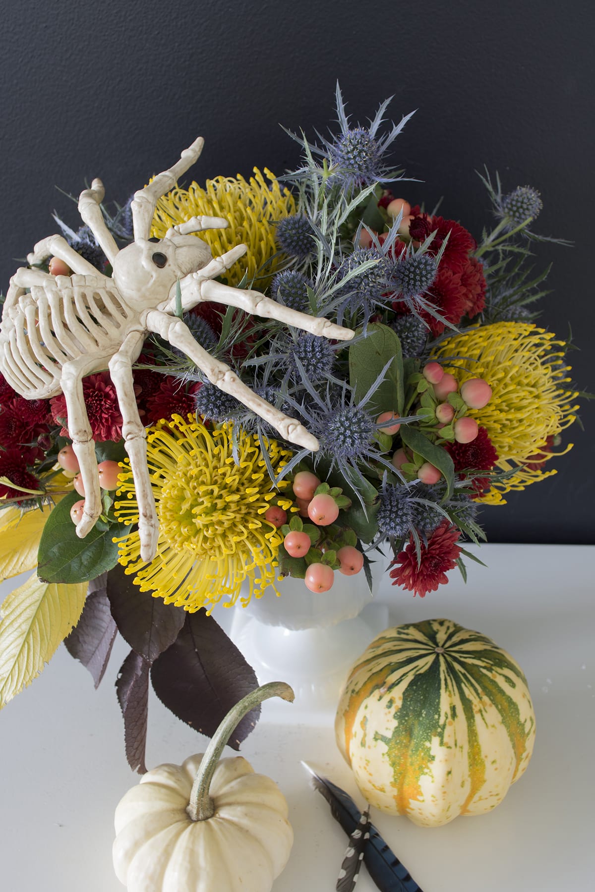 skeleton spider on top of vibrant colored fall arrangement with maroon chock cherry leaves, berries, dark red mums and pumkins with added larger skeleton spider for vibrant colored fall arrangement with maroon chock cherry leaves, berries, dark red mums and pumkins