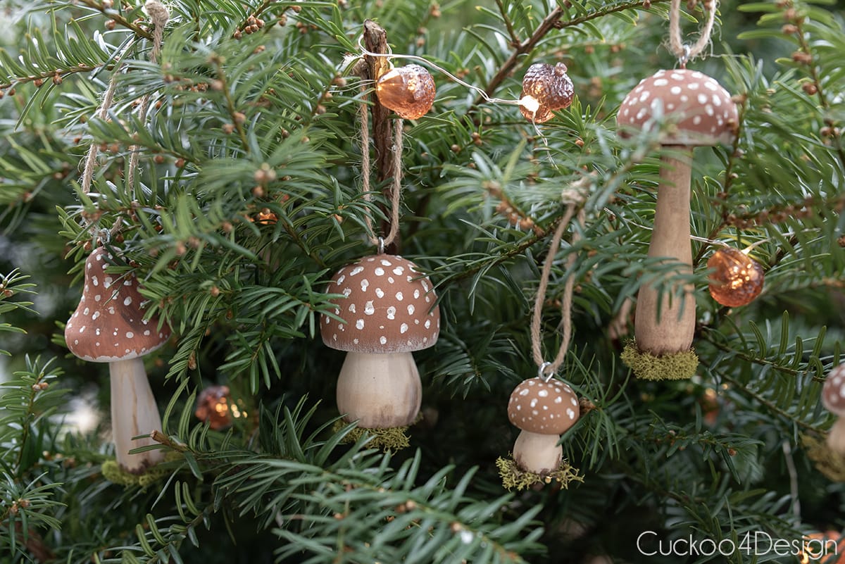 four mushroom Christmas ornaments with jute yarn twine hanging in pine tree with ahorn fairy lights