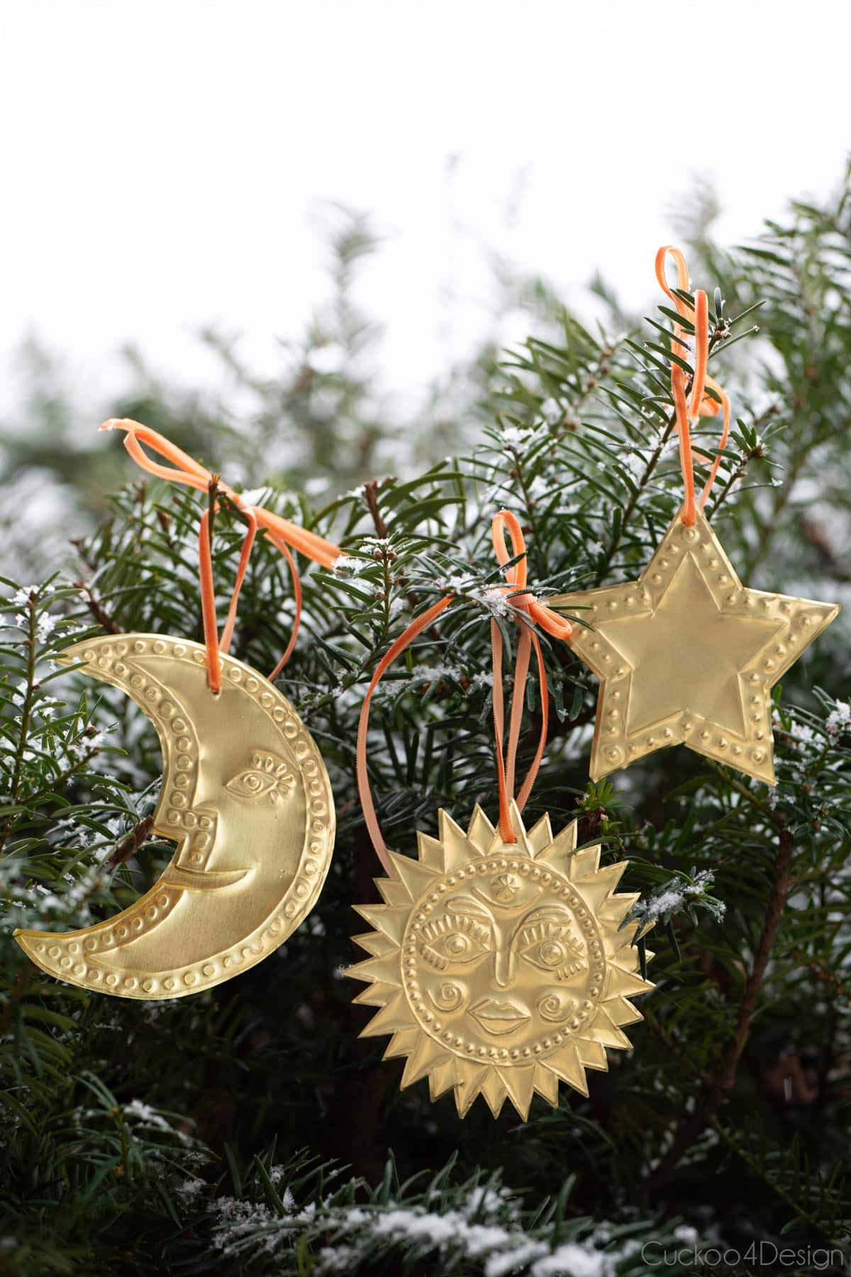 three different embossed tin ornaments hanging in a snowy pine tree