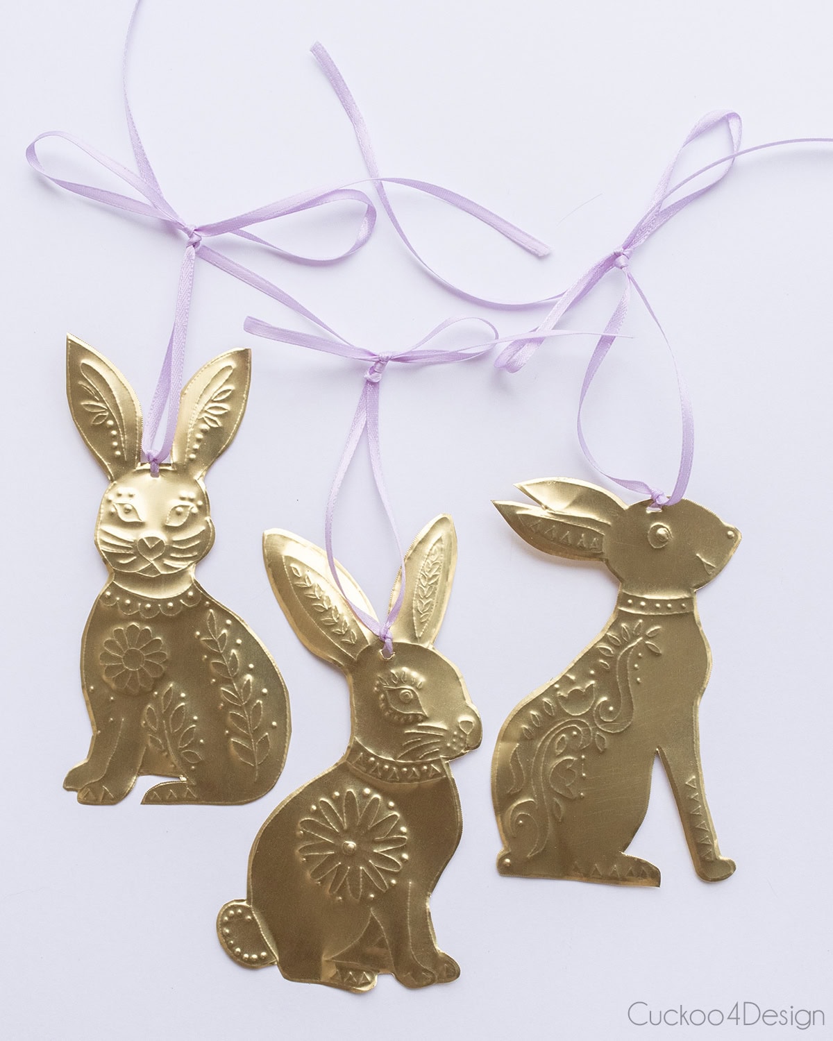 three brass Easter bunny ornaments with embossed folk art pattern and purple ribbons on white background