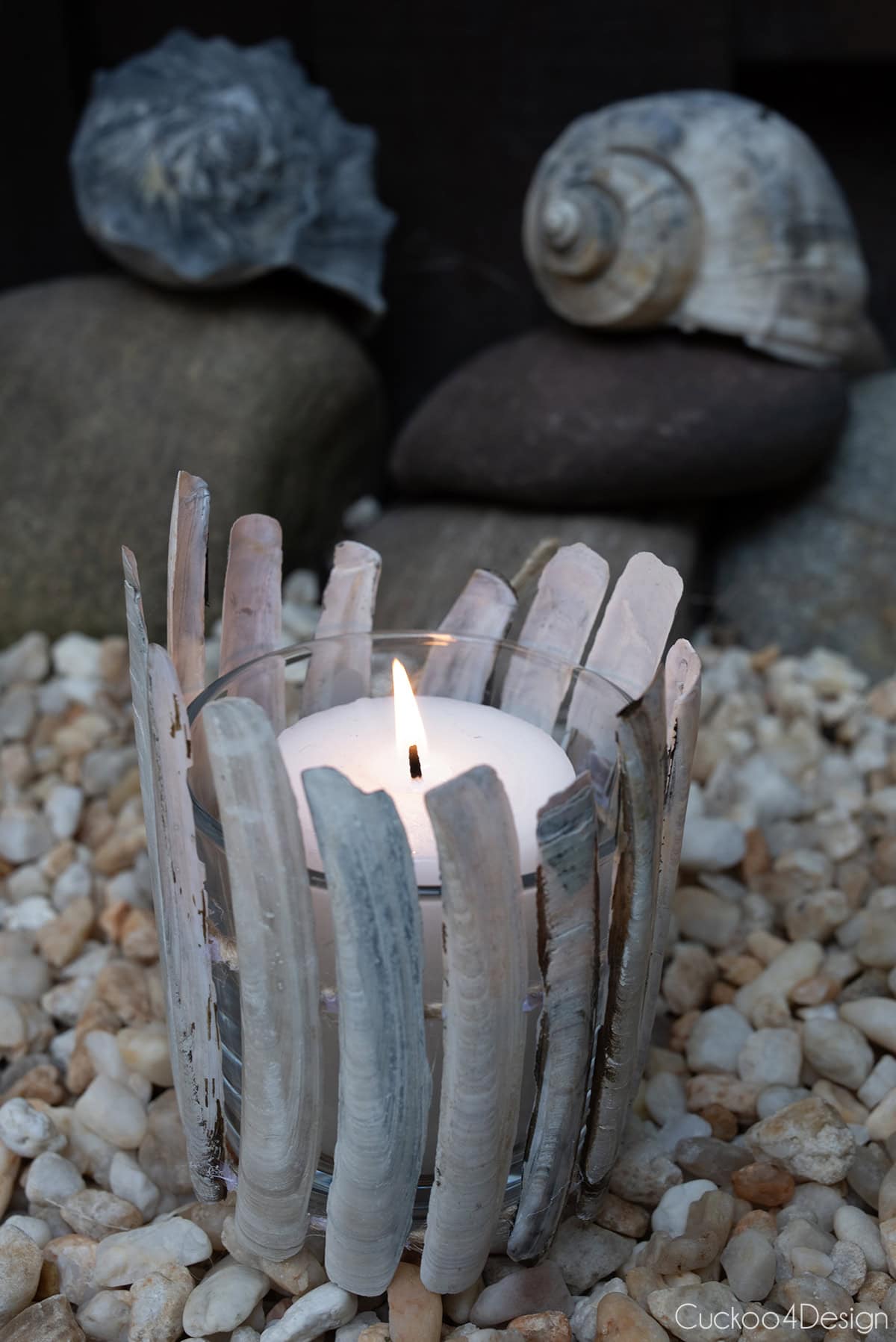 DIY seashell candle holder using razor clamshells on jute yarn, with lit candle, surrounded by beach pebbles and conch shells in the background