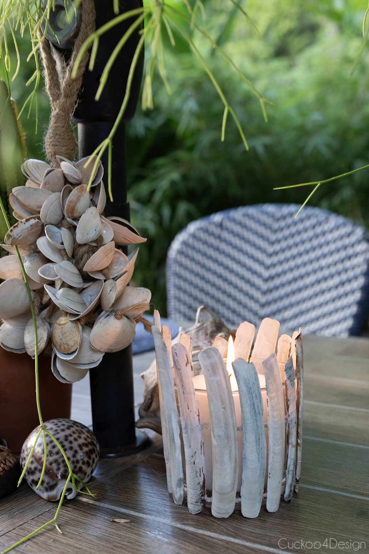 seashell candle holder using razor clamshells on jute yarn, with a lit candle, sitting on a patio table with a seashell tassel