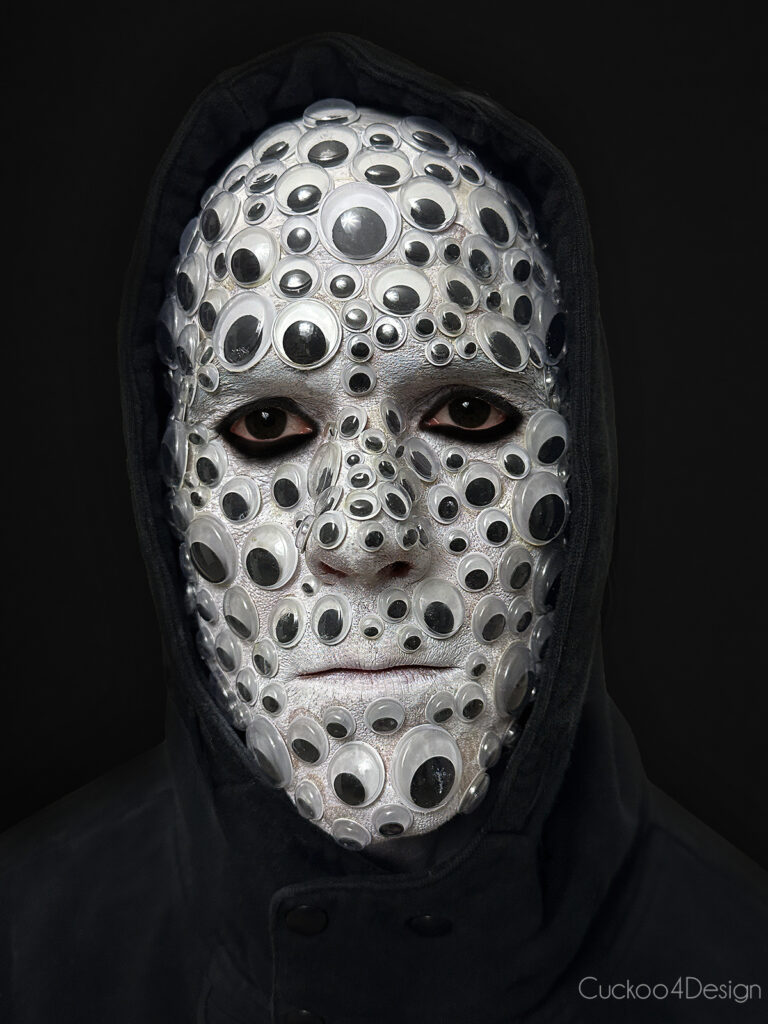 a man wearing black outfit and a full face of googly eyes glued to the face with black contact lenses