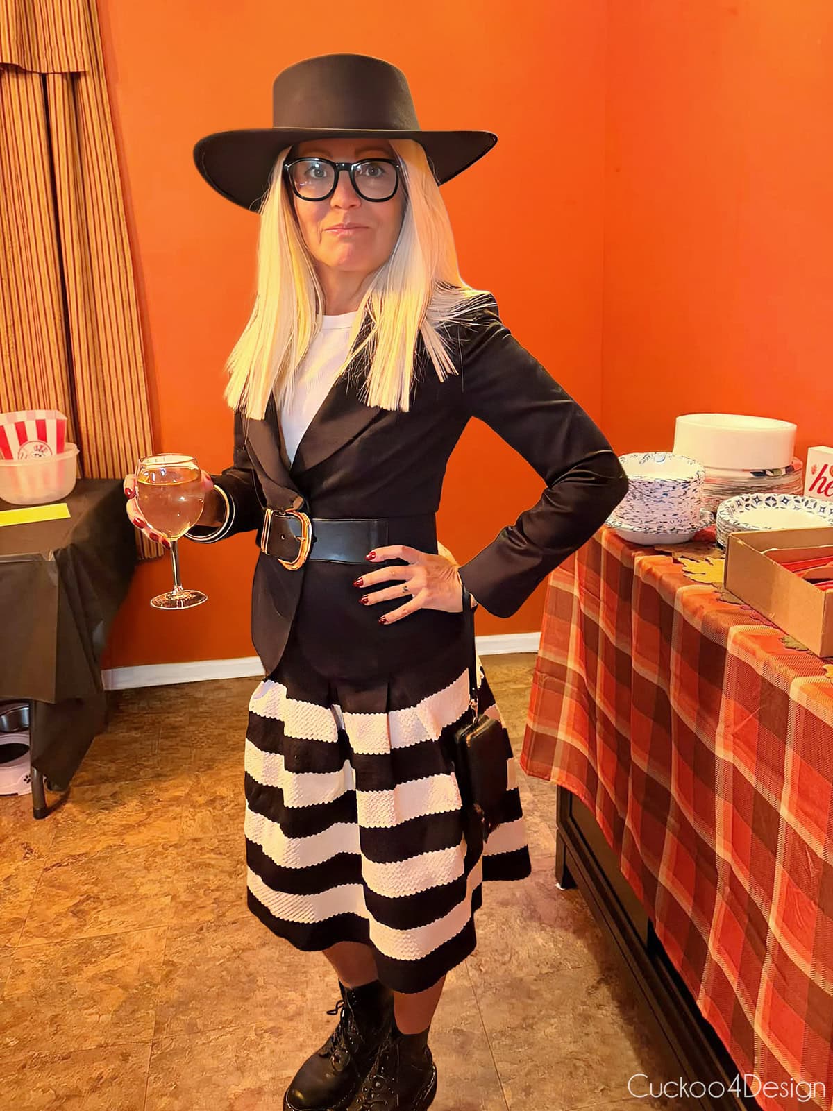 full length view of a Diane Keaton costume with a black hat, glasses, black blazer, black and white striped skirt, and black boots in front of orange background