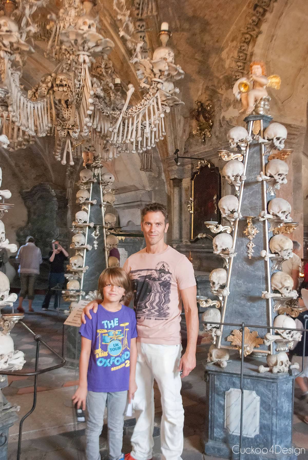 Father and son standing in front of a skull candelabra and the bone chandelier in the Sedlec Ossuary