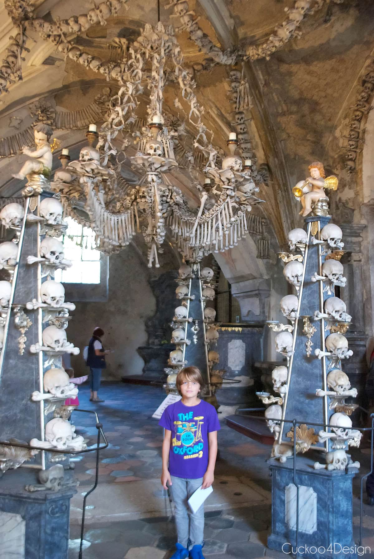 Boy standing between skull-covered candelabras under the bone chandelier in the Sedlec Ossuary